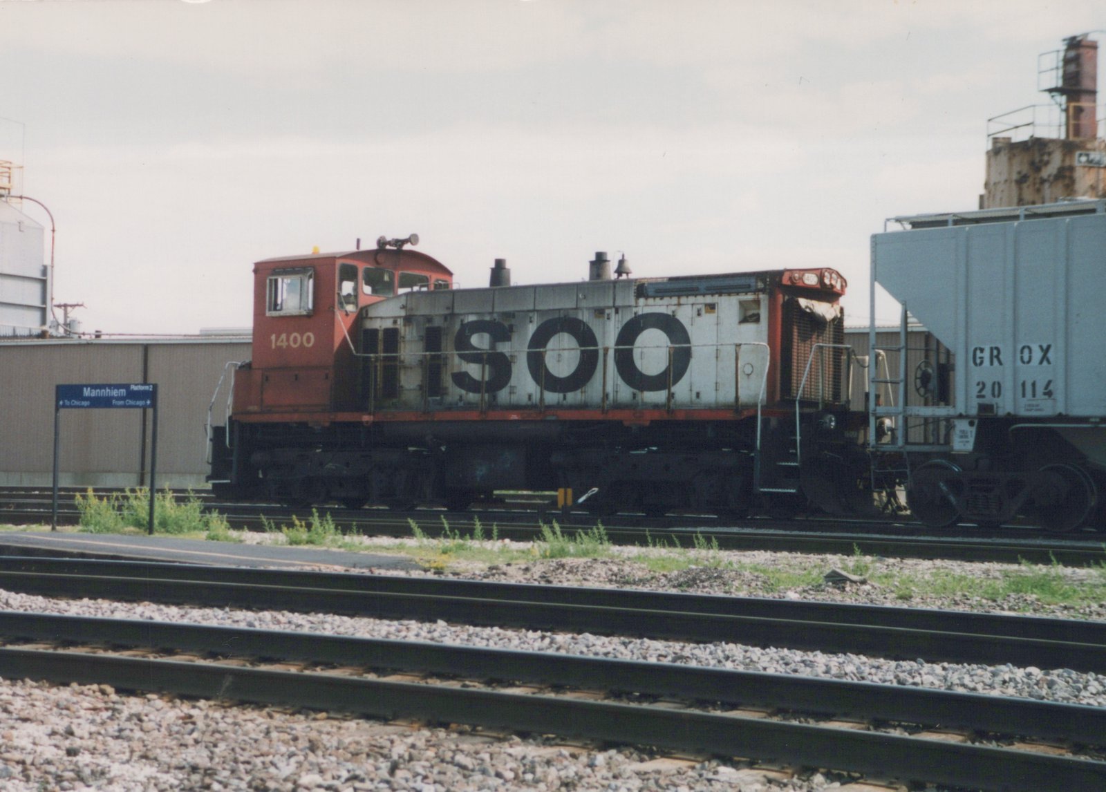 A side view of a red and grey SOO Line diesel locomotive numbered 1400, stopped on a track next to a station sign that reads 'Mannhiem Platform 2'.