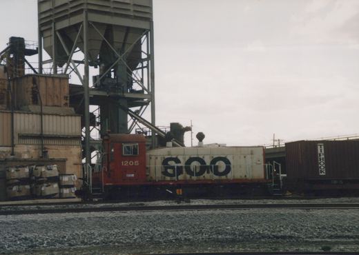 A side view of a red and white SOO Line diesel switcher locomotive, number 1205, on a railroad track next to a large industrial facility, with a dark brown boxcar labeled 'TIPHOOK' behind it.