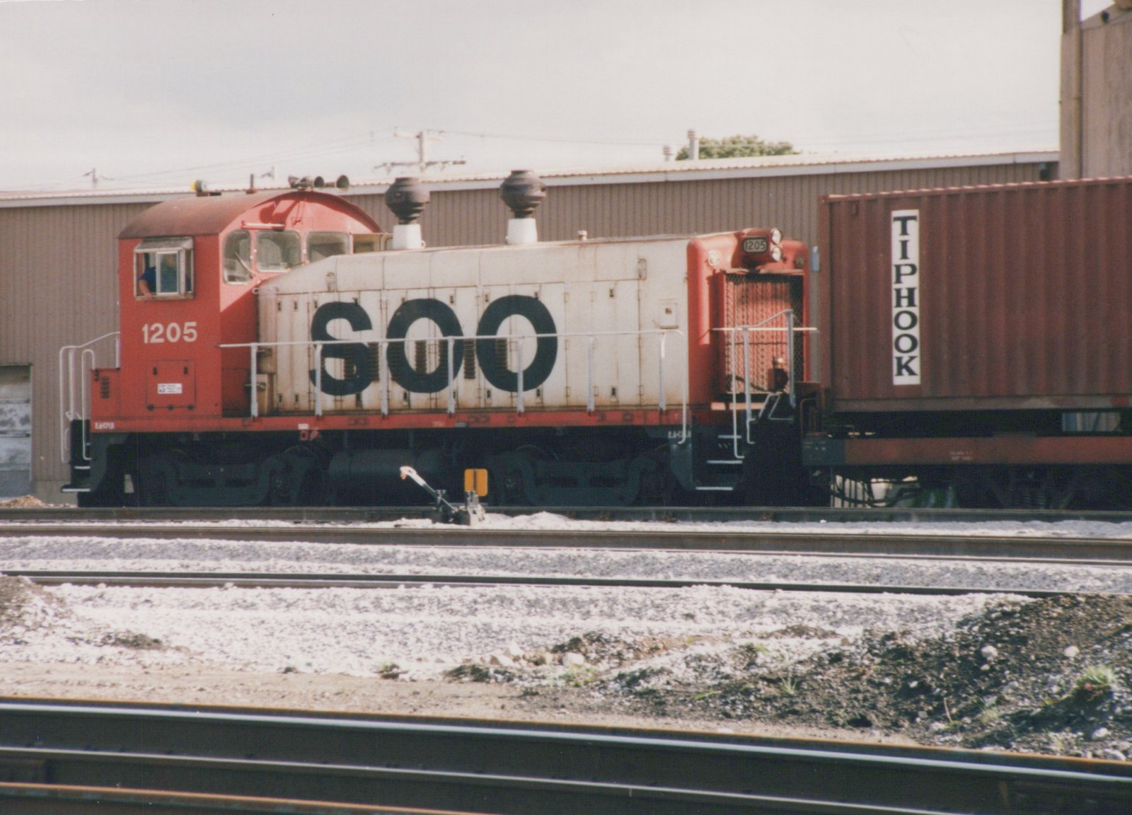 A red and white Soo Line diesel locomotive numbered 1205 is coupled to a flatcar with a brown Tiphook container, stopped on tracks in a gravel yard with a building behind it.