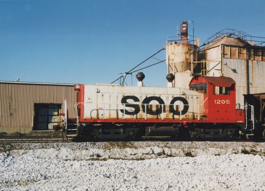 Side view of a red and white Soo Line diesel locomotive, number 1205, on a snowy track with industrial buildings in the background.