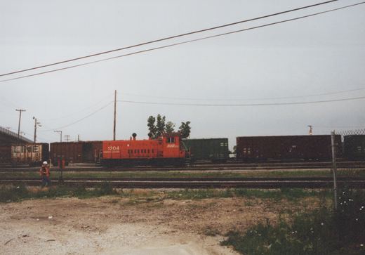 An orange SOO Line diesel locomotive numbered 1204 is parked on tracks in a freight yard, surrounded by various freight cars, including a brown Chicago, Rock Island and Pacific boxcar, under a cloudy sky.