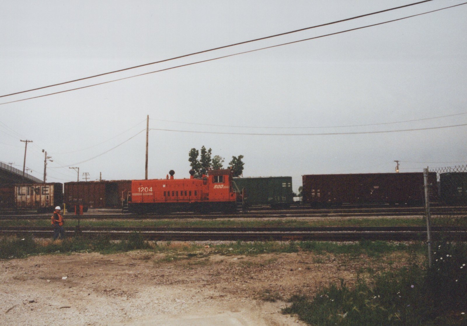 An orange SOO Line diesel locomotive numbered 1204 is parked on tracks in a freight yard, surrounded by various freight cars, including a brown Chicago, Rock Island and Pacific boxcar, under a cloudy sky.
