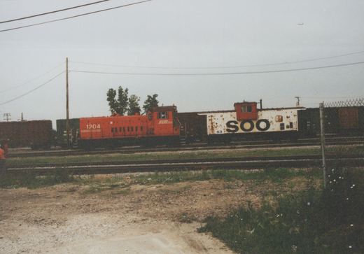 A red SOO Line diesel locomotive numbered 1204 pulls a freight train with a white SOO caboose numbered 63 along railroad tracks, with a person standing nearby on a gravelly field under an overcast sky.