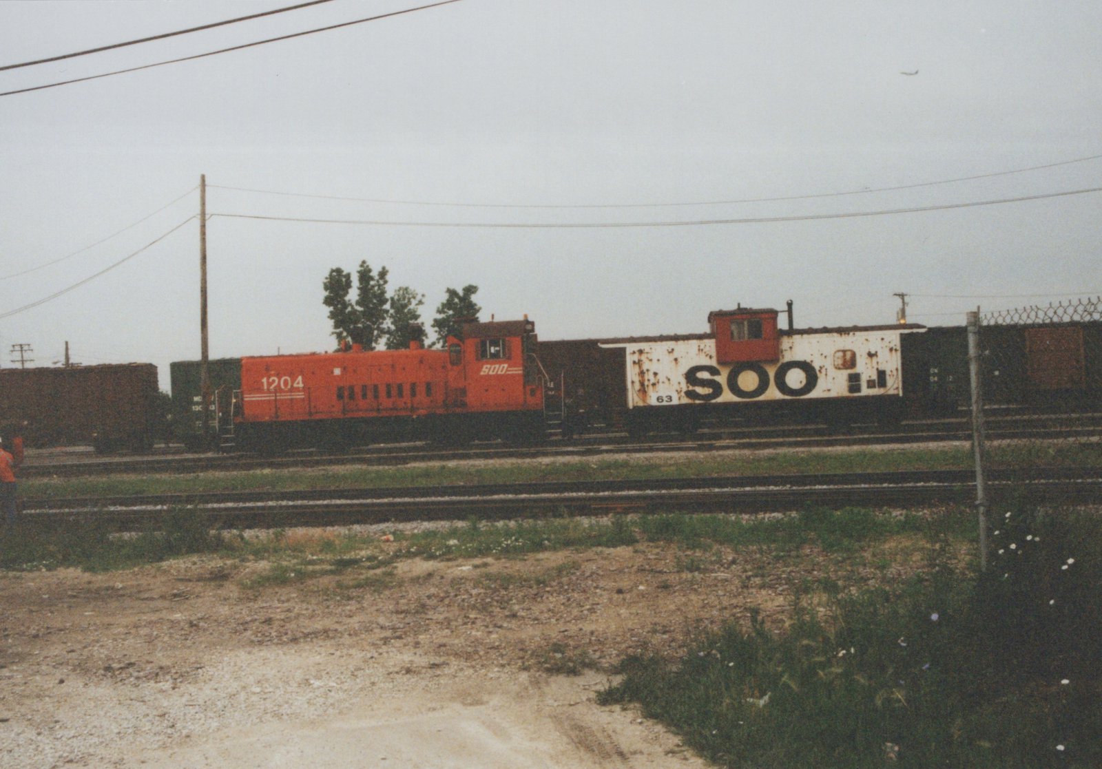 A red SOO Line diesel locomotive numbered 1204 pulls a freight train with a white SOO caboose numbered 63 along railroad tracks, with a person standing nearby on a gravelly field under an overcast sky.