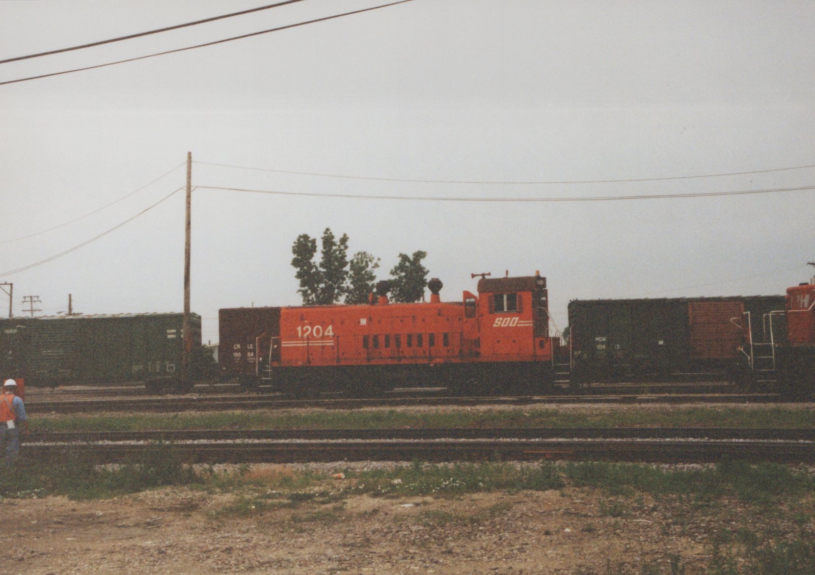 An orange Soo Line EMD SW1200 diesel locomotive numbered 1204 sits on tracks in a yard, flanked by dark green boxcars from the Chicago and North Western and Milwaukee Road railroads, under an overcast sky.