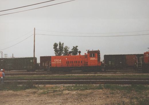 An orange Soo Line EMD SW1200 diesel locomotive numbered 1204 sits on tracks in a yard, flanked by dark green boxcars from the Chicago and North Western and Milwaukee Road railroads, under an overcast sky.