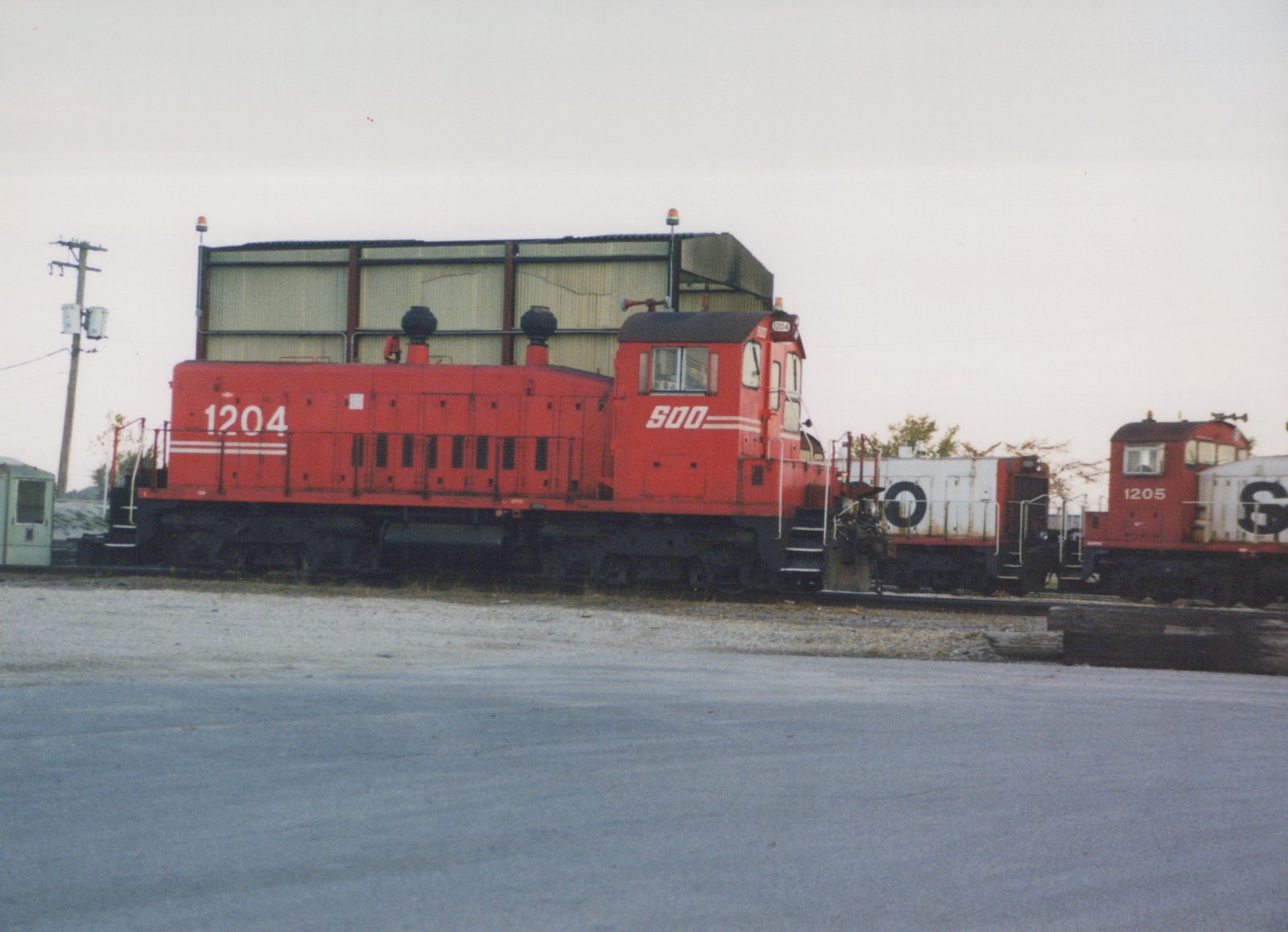 A red Soo Line diesel locomotive numbered 1204 is parked on tracks in a gravel yard, with another red locomotive and a white boxcar behind it under an overcast sky.