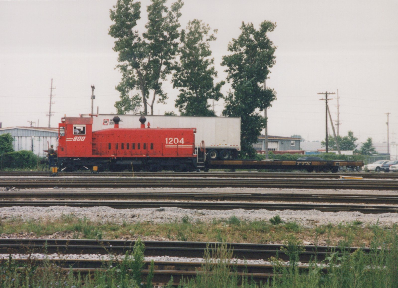 A red SOO Line locomotive number 1204 stands on multiple tracks in a rail yard, pulling a flatcar with a trailer. Two people are visible on the locomotive.
