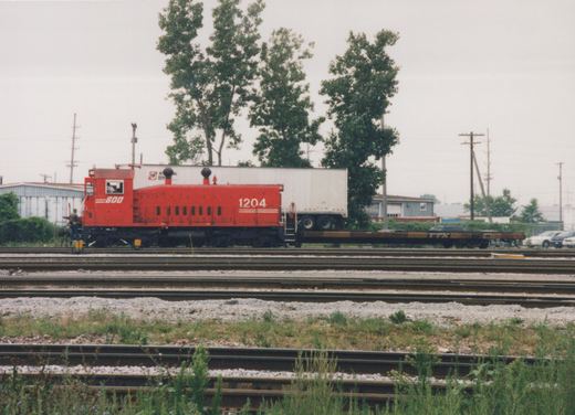 A red SOO Line locomotive number 1204 stands on multiple tracks in a rail yard, pulling a flatcar with a trailer. Two people are visible on the locomotive.