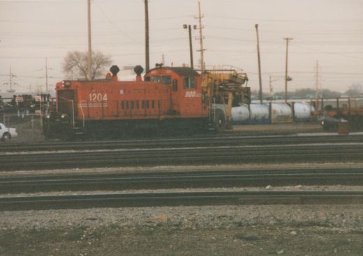 An orange SOO Line diesel locomotive with the number 1204 is parked on a set of tracks in a yard, with other rail cars and equipment in the background.