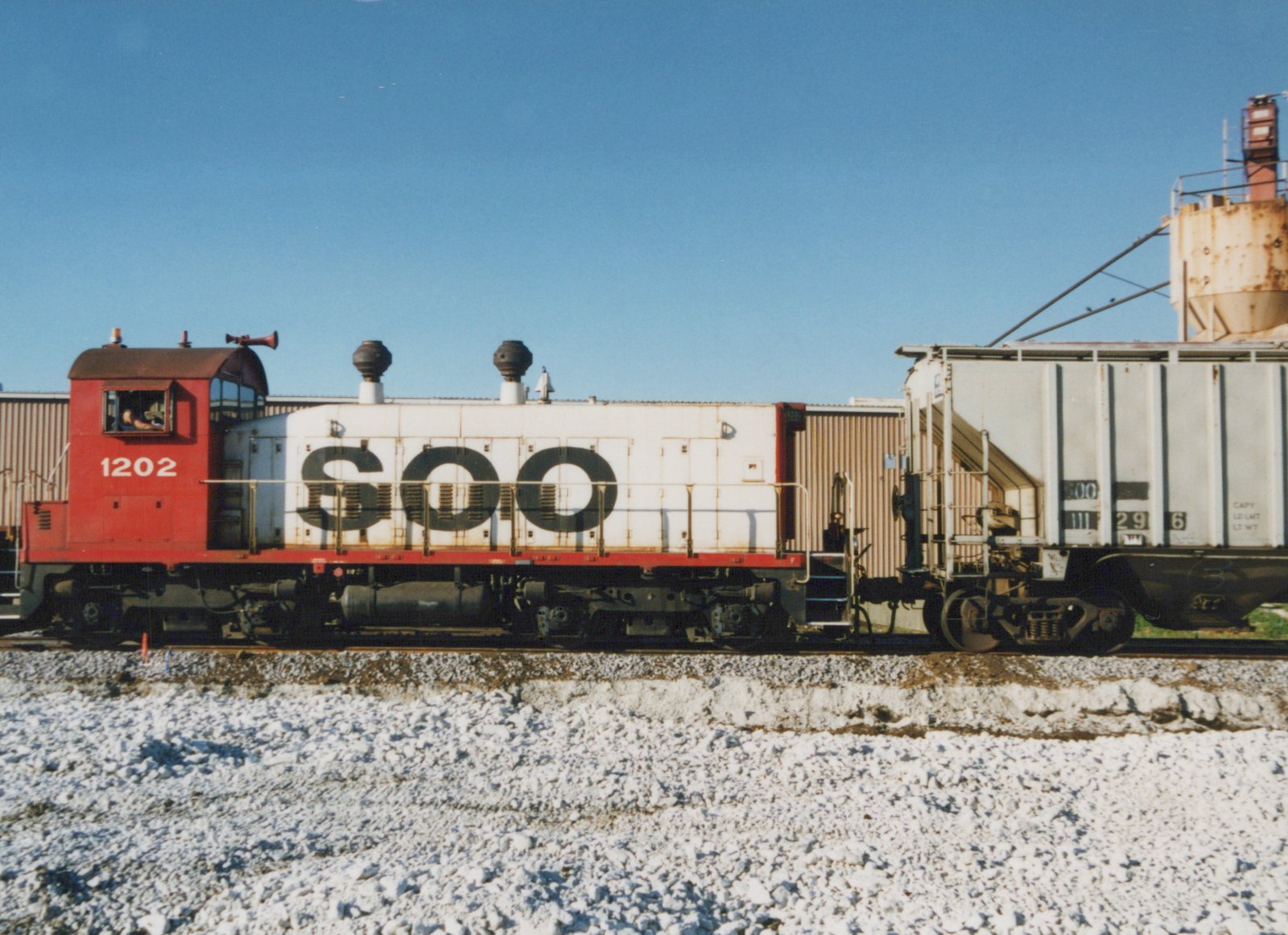 Side view of a red and white Soo Line EMD SW1200 diesel locomotive numbered 1202, coupled to a freight car, in a snowy yard under a clear blue sky.