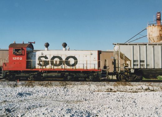 Side view of a red and white Soo Line EMD SW1200 diesel locomotive numbered 1202, coupled to a freight car, in a snowy yard under a clear blue sky.