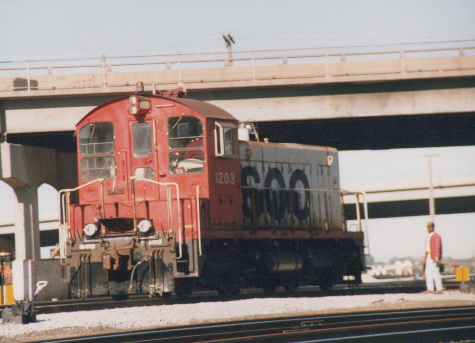 A red and gray SOO Line diesel locomotive numbered 1203 is positioned on train tracks beneath a bridge, with one person standing nearby.