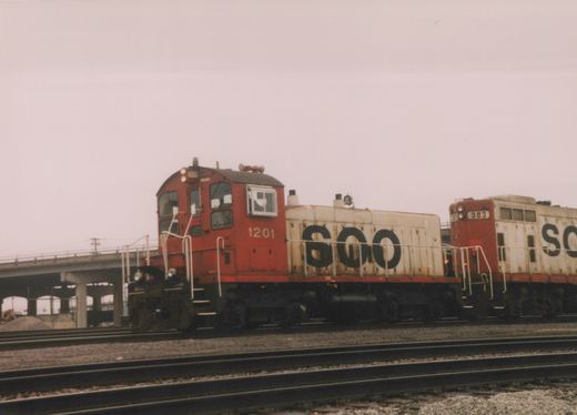 Two red and white SOO Line diesel locomotives, numbered 1201 and 383, are parked on parallel tracks beside a concrete bridge on an overcast day.