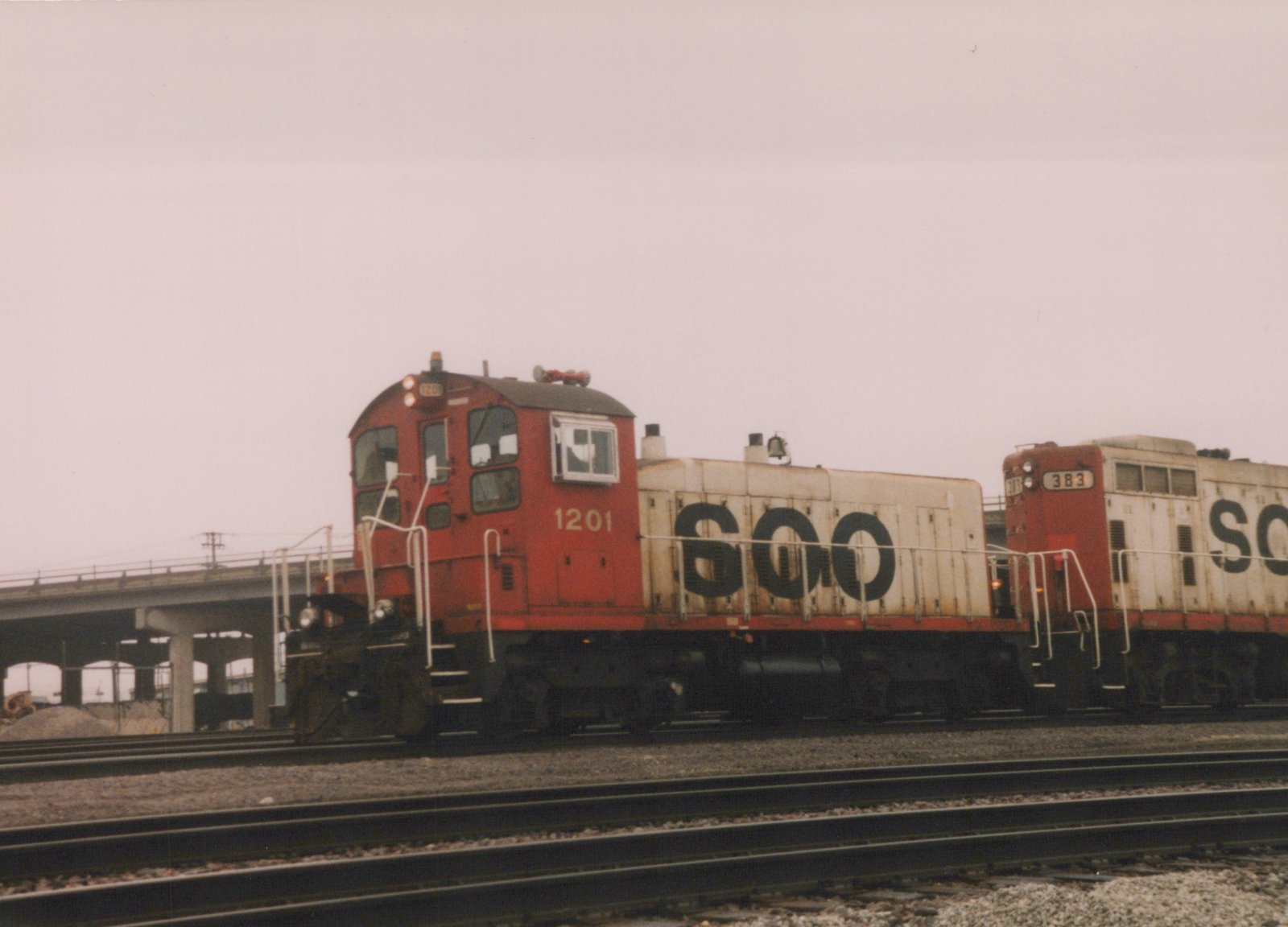 Two red and white SOO Line diesel locomotives, numbered 1201 and 383, are parked on parallel tracks beside a concrete bridge on an overcast day.