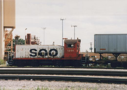 Side view of a red and white Soo Line diesel locomotive numbered 1201, standing on tracks next to a flatcar carrying a semi-trailer, with industrial buildings and utility poles in the background under an overcast sky.