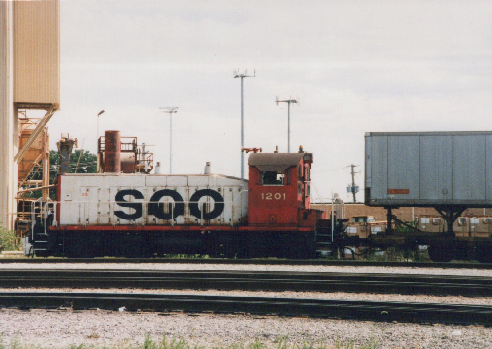 Side view of a red and white Soo Line diesel locomotive numbered 1201, standing on tracks next to a flatcar carrying a semi-trailer, with industrial buildings and utility poles in the background under an overcast sky.
