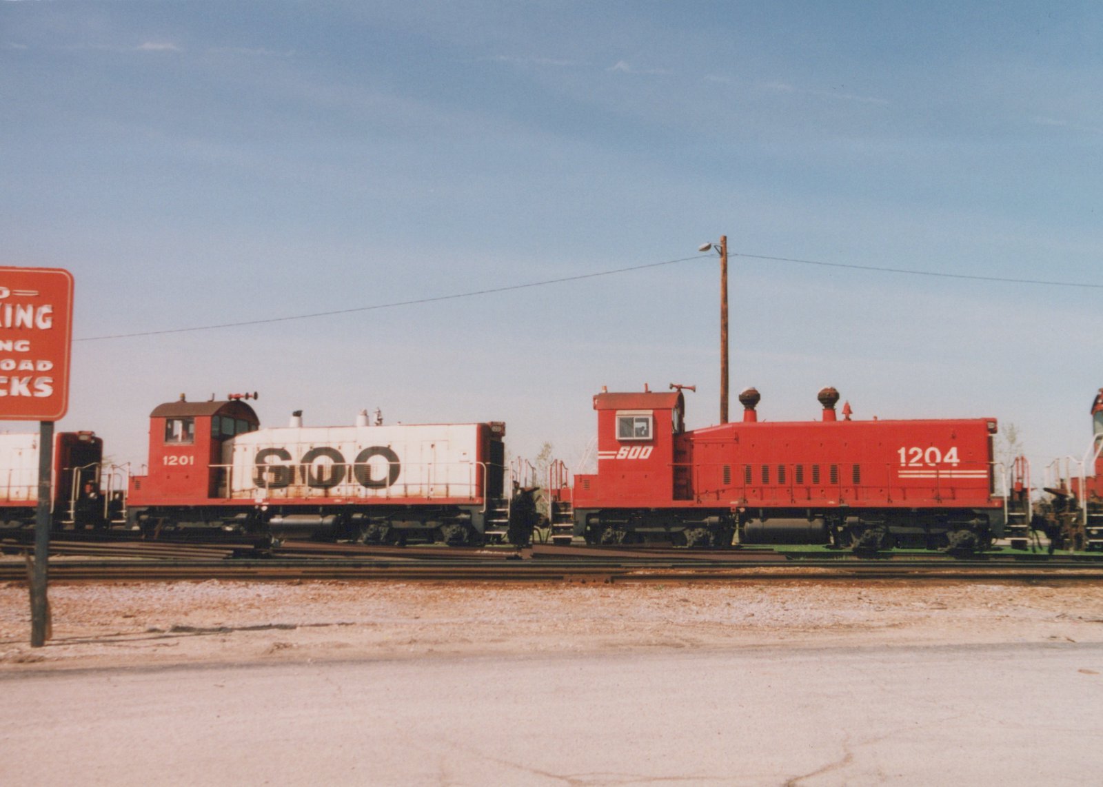 Two red Soo Line diesel locomotives, numbered 1201 and 1204, are parked on parallel tracks in a gravel rail yard on a sunny day, with a road sign partially visible on the left.
