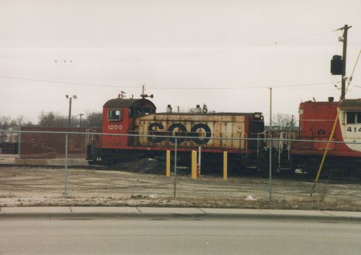 A red and cream SOO Line locomotive number 1200 is parked behind a chain-link fence in a snowy yard, with another locomotive partially visible.