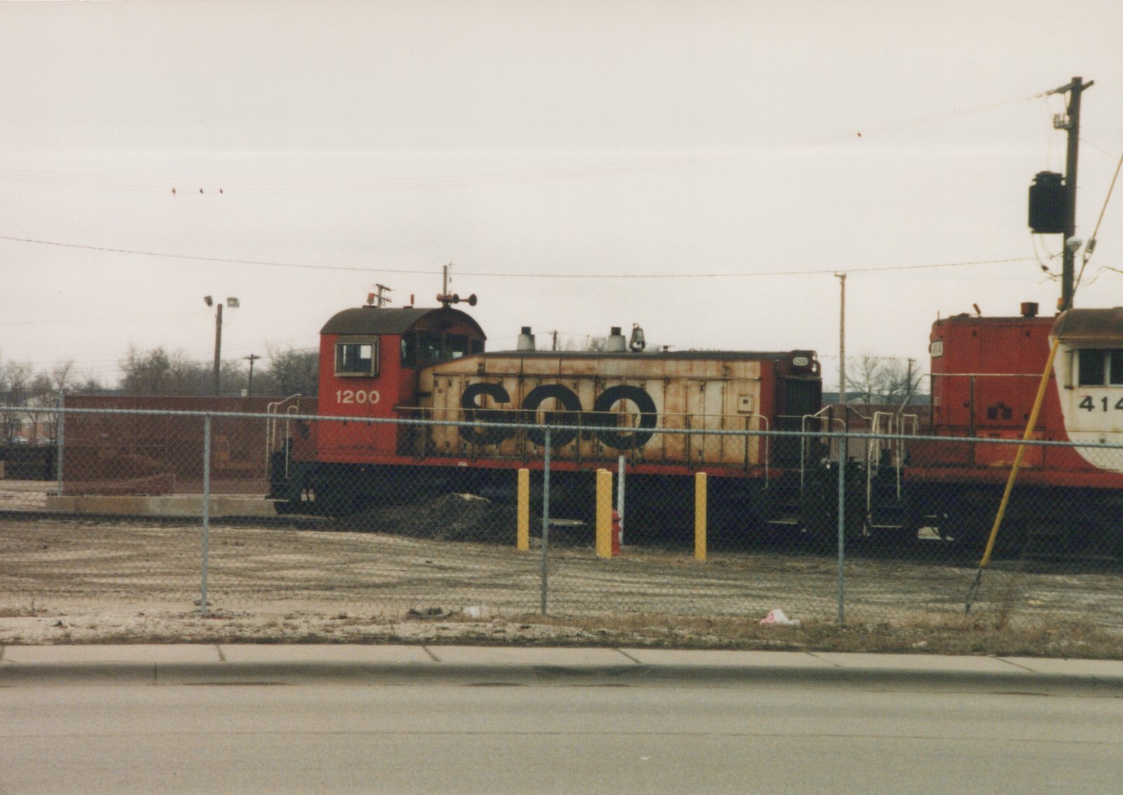 A red and cream SOO Line locomotive number 1200 is parked behind a chain-link fence in a snowy yard, with another locomotive partially visible.