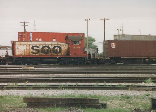 A red and cream SOO Line caboose numbered 1200 sits on a track in a rail yard, with orange and brown shipping containers on flatcars behind it under an overcast sky.