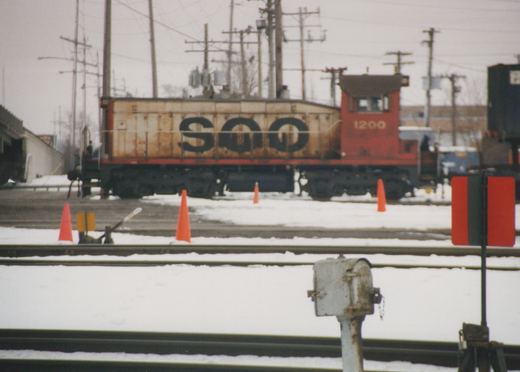 A rusty brown and red Soo Line locomotive, number 1200, is parked on snow-covered tracks in a rail yard.