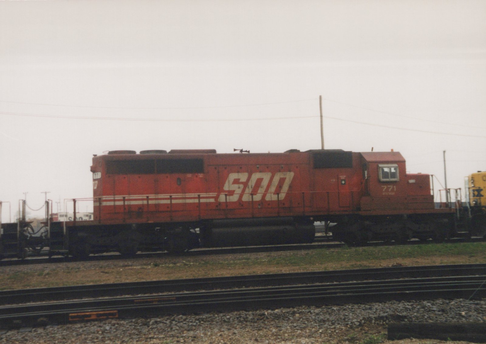 A red SOO Line diesel locomotive with the number 771 on its side, parked on tracks in a gravel yard under a gray sky.