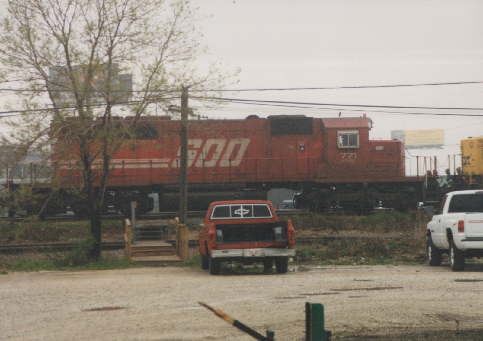 A red SOO Line diesel locomotive numbered 771 is seen at a grade crossing with an orange pickup truck and a white pickup truck parked nearby, under an overcast sky.