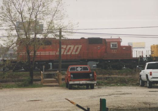 A red SOO Line diesel locomotive numbered 771 is seen at a grade crossing with an orange pickup truck and a white pickup truck parked nearby, under an overcast sky.