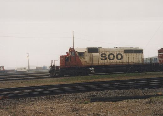 A side view of a Soo Line diesel locomotive numbered 775, with a crew member standing on the platform, in a rail yard under an overcast sky.