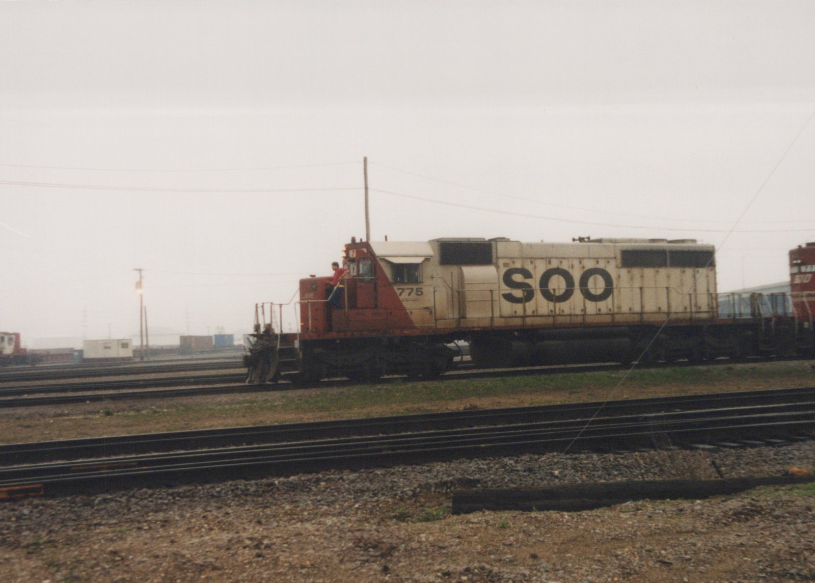 A side view of a Soo Line diesel locomotive numbered 775, with a crew member standing on the platform, in a rail yard under an overcast sky.