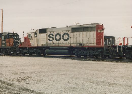 A side view of a Soo Line diesel locomotive numbered 763, painted white with red ends, standing on tracks in a gravel yard with another locomotive behind it.