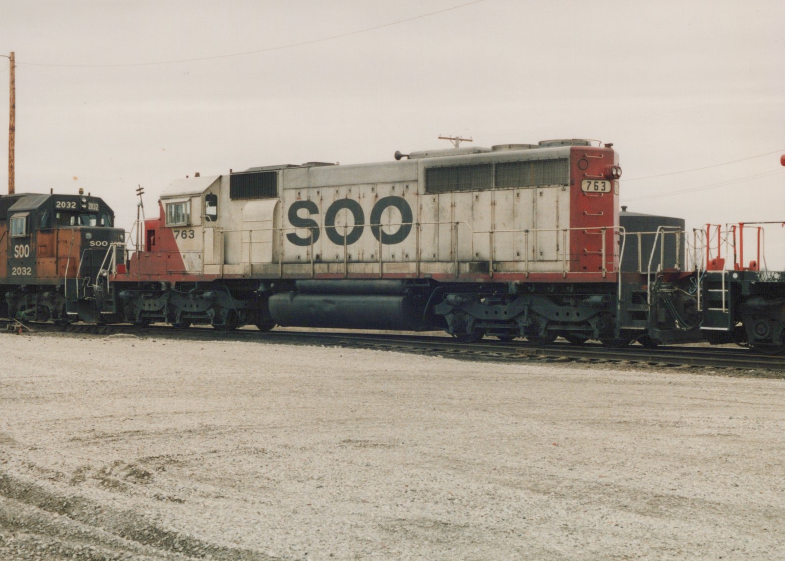 A side view of a Soo Line diesel locomotive numbered 763, painted white with red ends, standing on tracks in a gravel yard with another locomotive behind it.