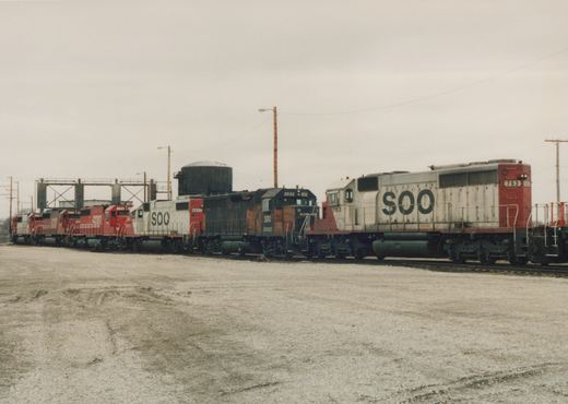 Multiple Soo Line locomotives, including numbers 763, 2032, 4437, and 755, are lined up on tracks in a gravel yard on a cloudy day.