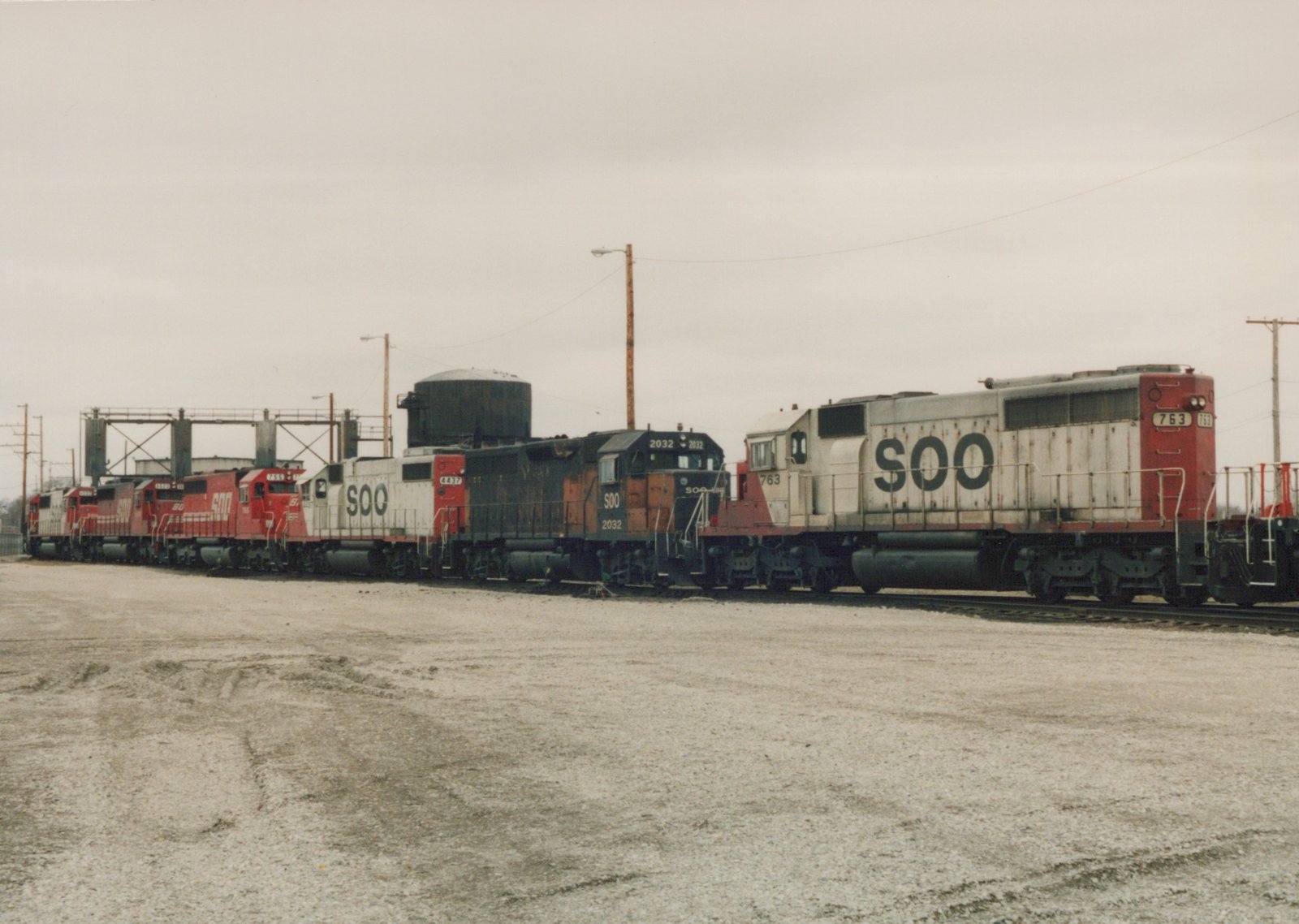 Multiple Soo Line locomotives, including numbers 763, 2032, 4437, and 755, are lined up on tracks in a gravel yard on a cloudy day.