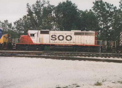 A white and red SOO Line diesel locomotive numbered 765 sits on railroad tracks in a yard, with trees in the background under an overcast sky.
