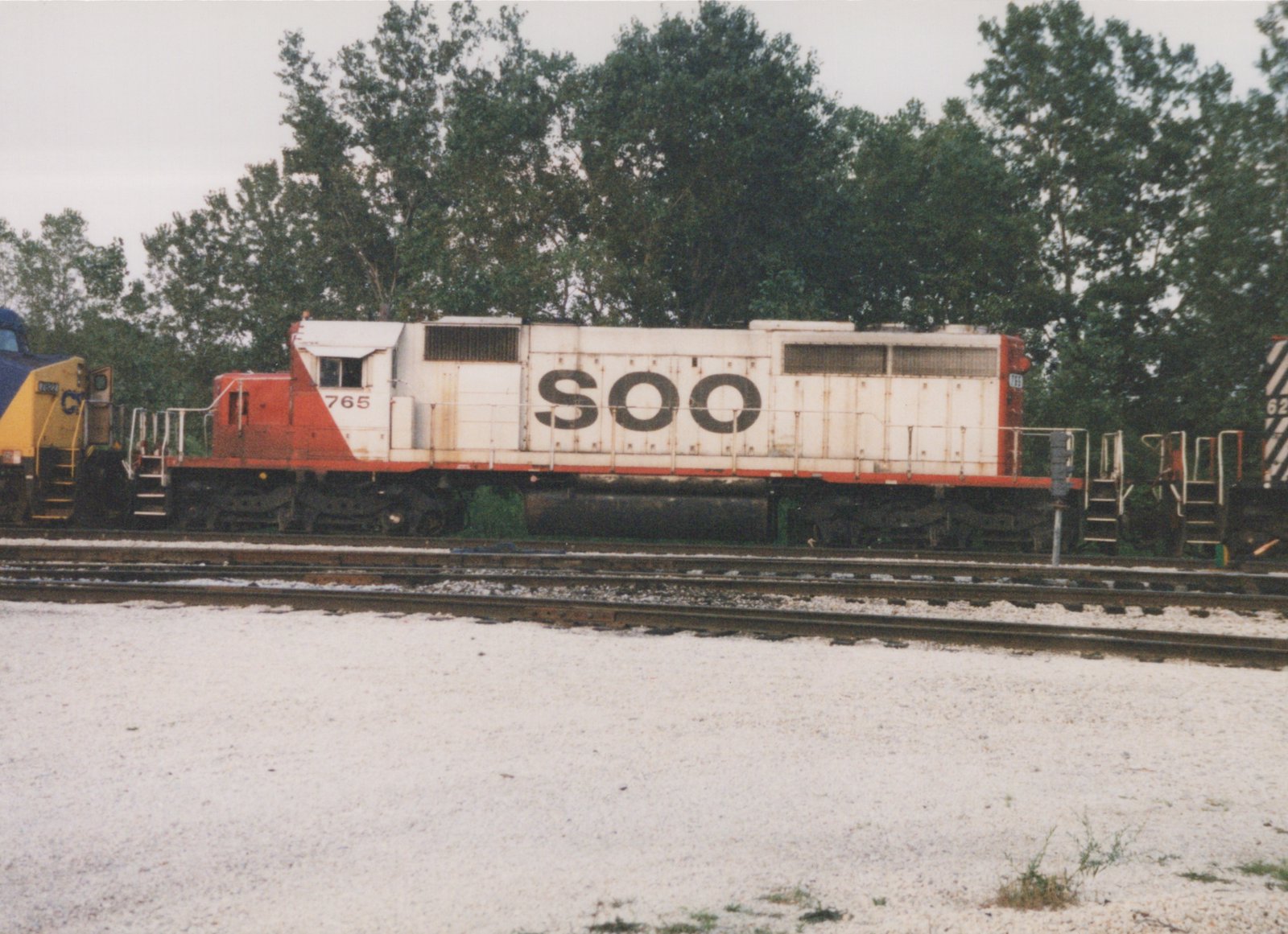 A white and red SOO Line diesel locomotive numbered 765 sits on railroad tracks in a yard, with trees in the background under an overcast sky.