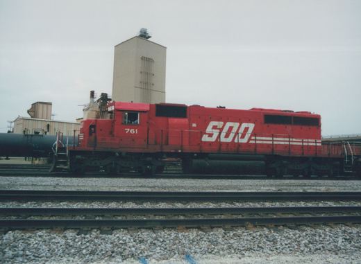 A red Soo Line locomotive numbered 761 is shown from the side, sitting on railroad tracks with an industrial building in the background under a gray sky.
