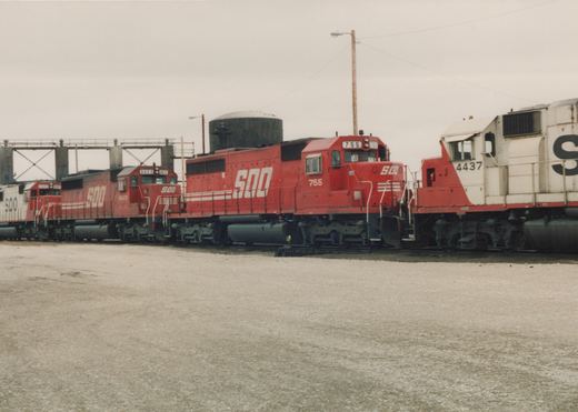 A line of three red Soo Line diesel locomotives parked in a gravel yard with a water tower and industrial structures in the background.