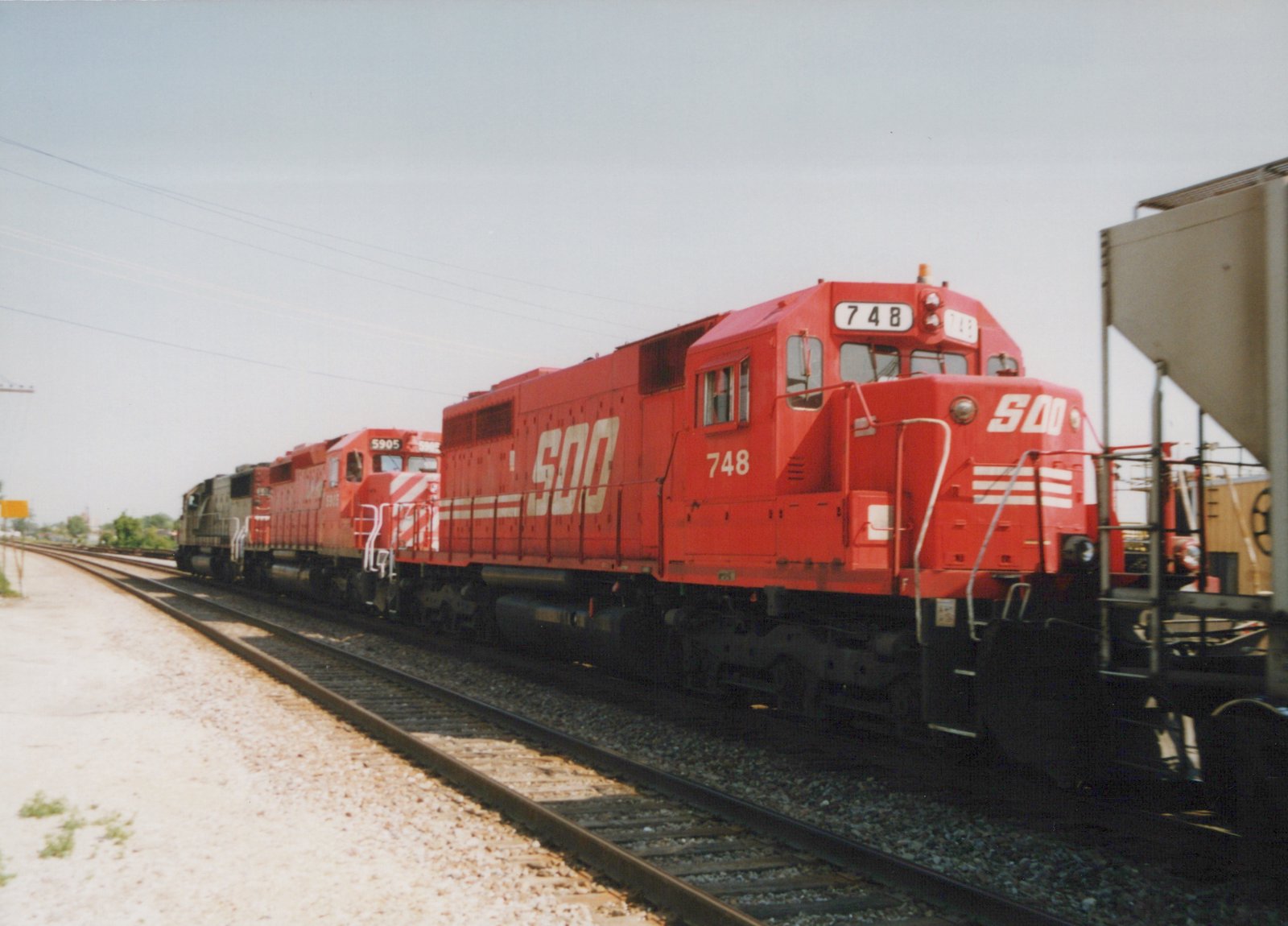 A three-unit consist of red SOO Line diesel locomotives, led by number 748, is coupled to freight cars on a railroad main line under an overcast sky.