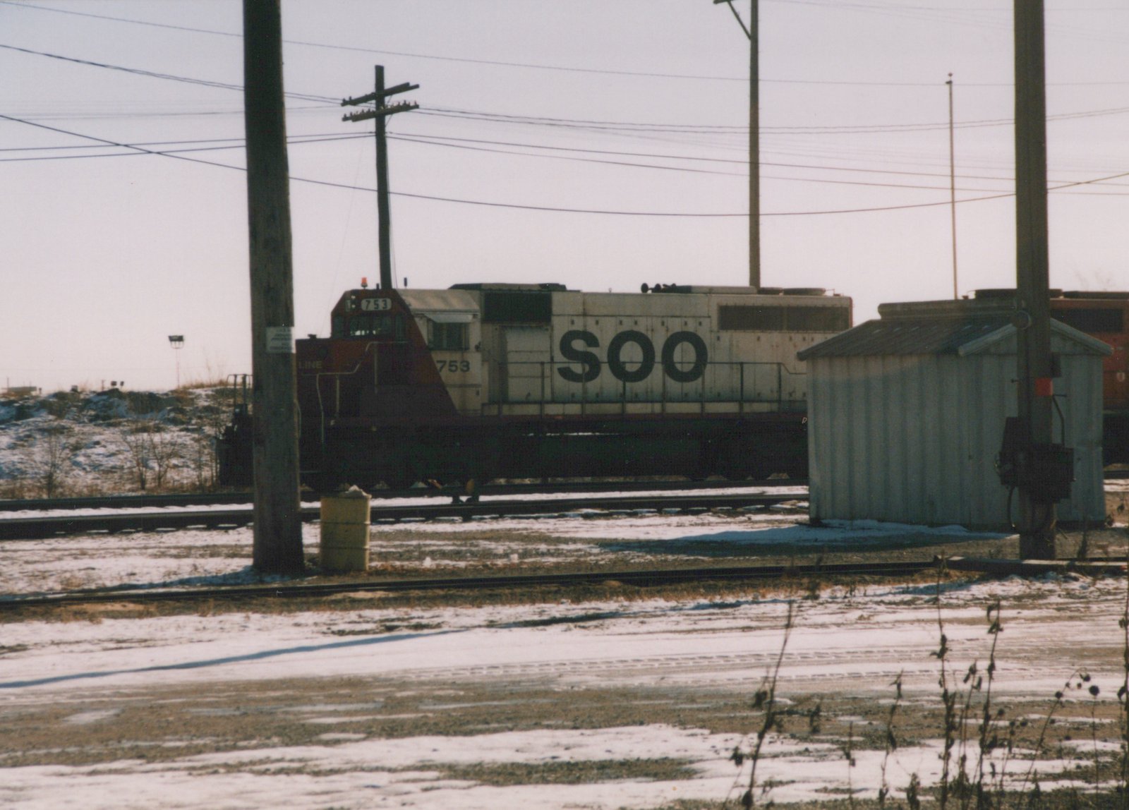 A white and maroon Soo Line locomotive numbered 753 is parked on a snow-covered railroad yard next to a small shed and utility poles.