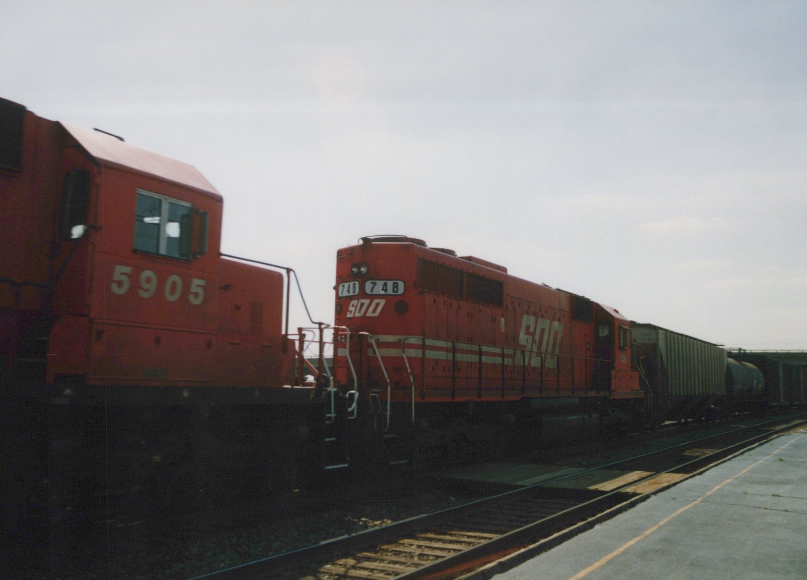 Two red diesel locomotives, one with the SOO Line logo and number 748, on parallel tracks in a yard under an overcast sky.