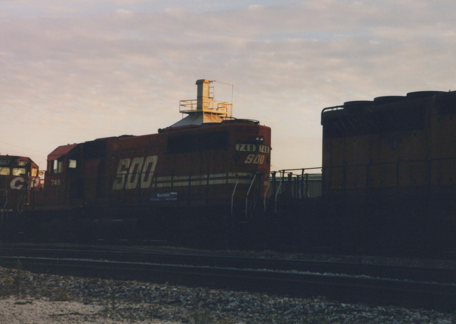 A red SOO Line diesel locomotive, number 748, is shown from a three-quarter front view in a railroad yard at dusk, with its side and front numbers clearly visible. A second locomotive with CP lettering is partially seen to the left.
