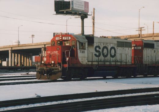 Two red and white SOO Line diesel locomotives on a snowy railroad yard, with a highway overpass and a billboard in the background.