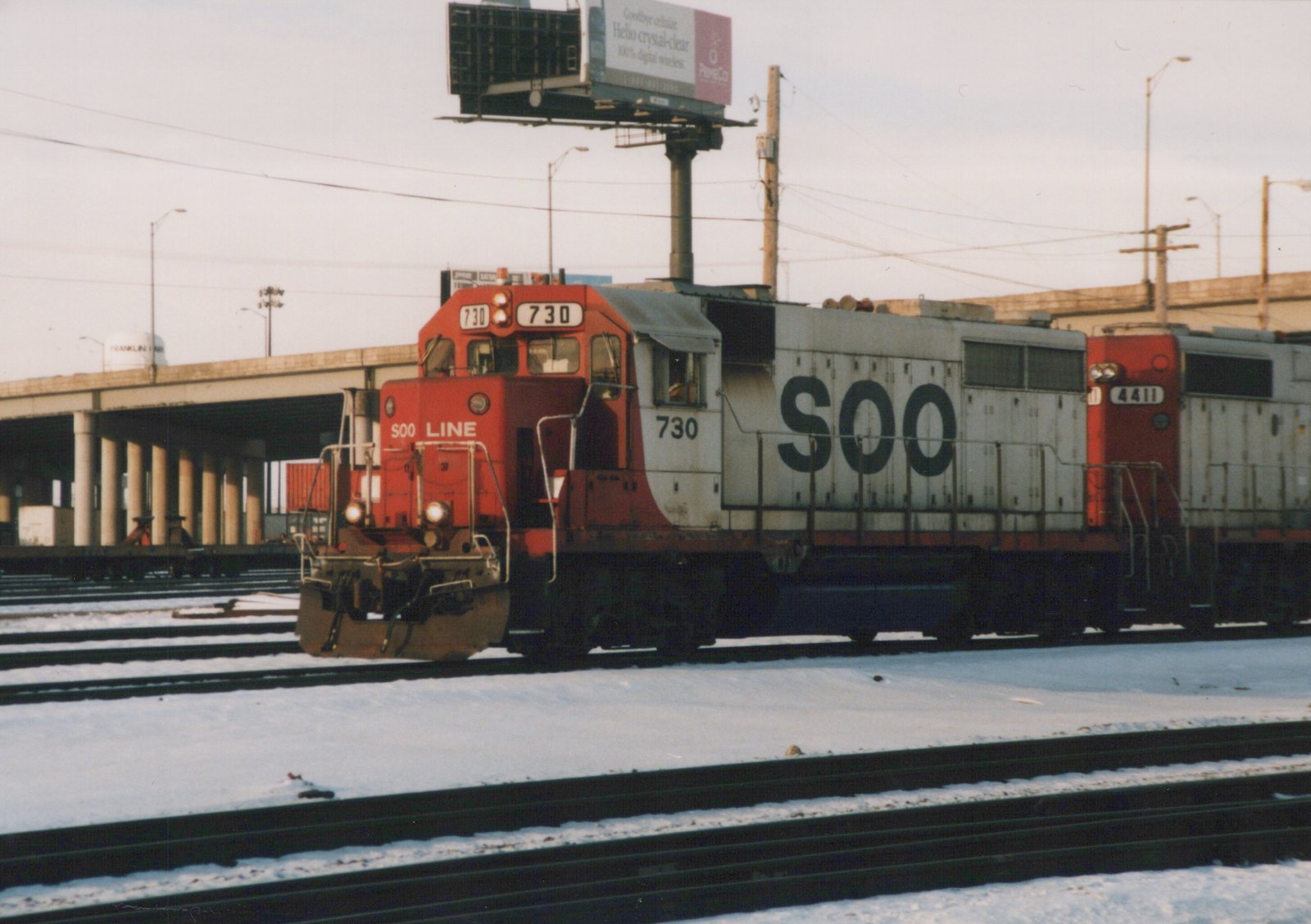 Two red and white SOO Line diesel locomotives on a snowy railroad yard, with a highway overpass and a billboard in the background.