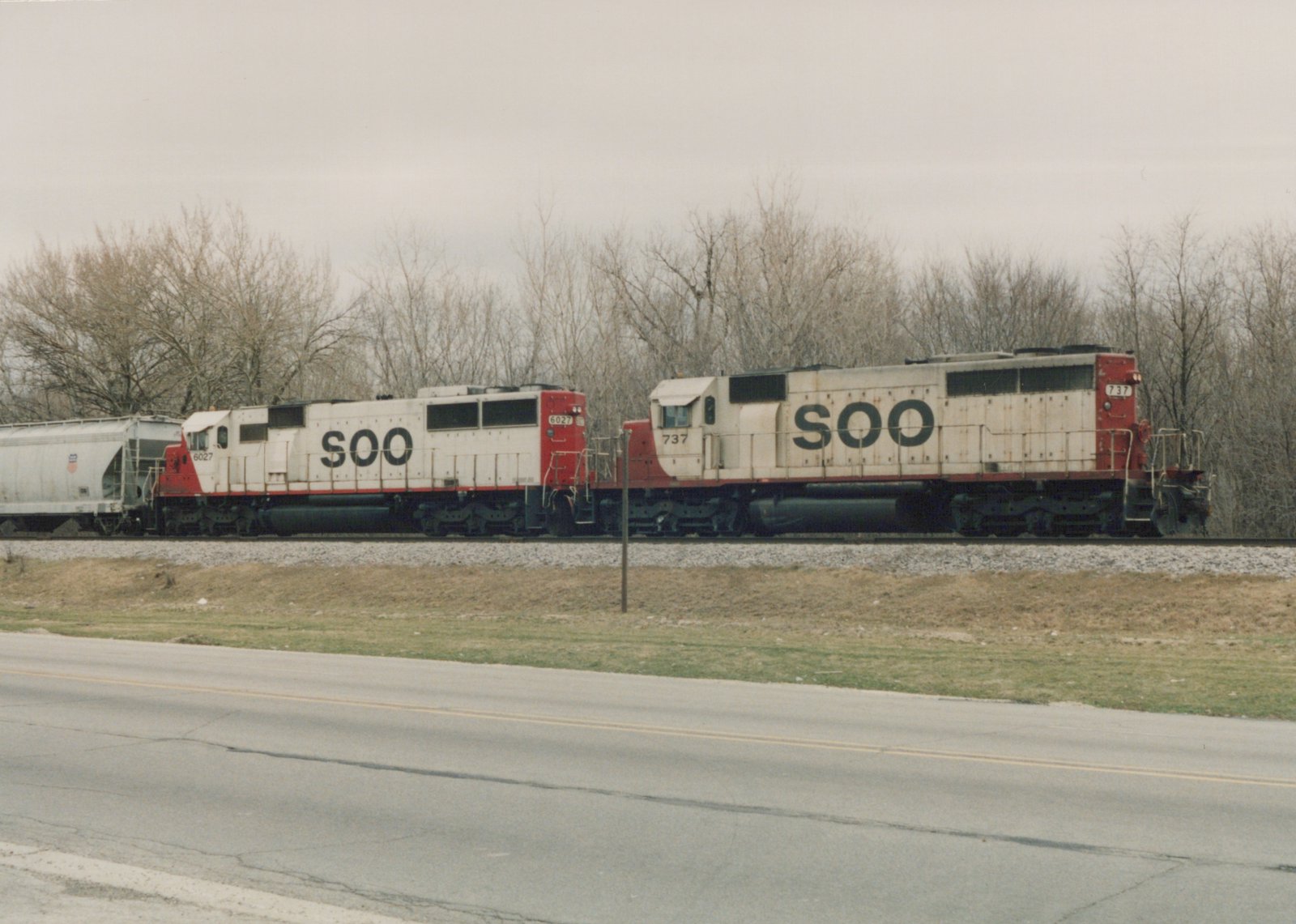 Side view of a Soo Line freight train with two locomotives, numbers 6027 and 737, on a railroad track beside a road with bare trees in the background.