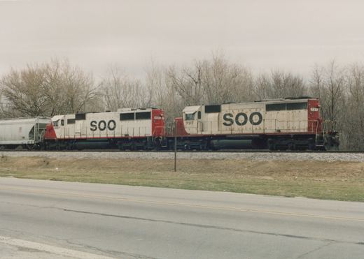 Side view of a Soo Line freight train with two locomotives, numbers 6027 and 737, on a railroad track beside a road with bare trees in the background.
