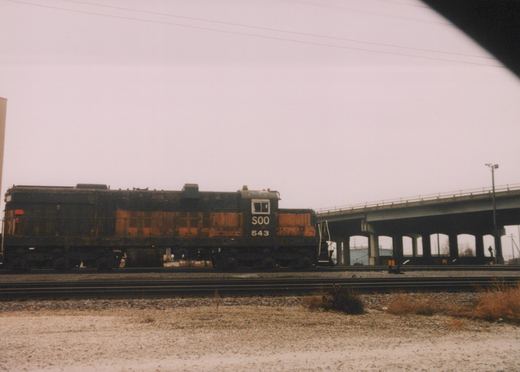 A side view of SOO Line locomotive 543, an EMD SW1200, standing on tracks beside a concrete overpass under an overcast sky.