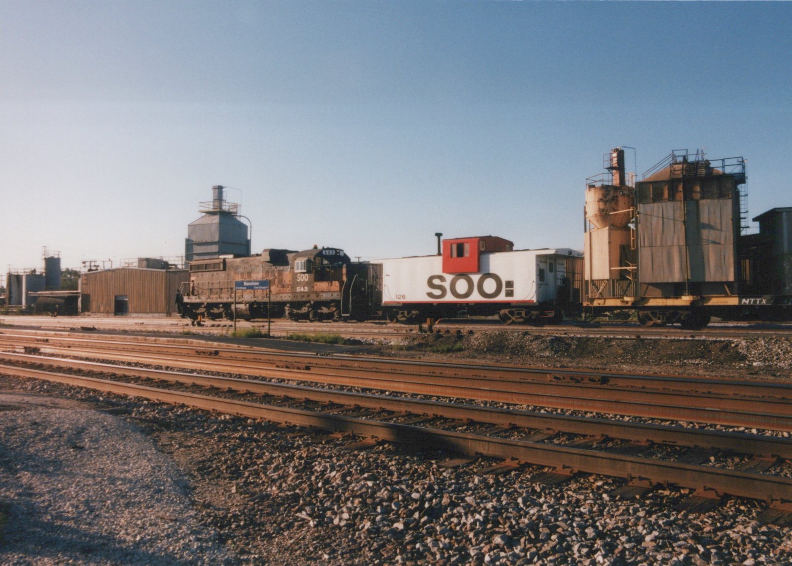 A SOO Line diesel locomotive and caboose are parked on parallel tracks in a sunny rail yard, with industrial buildings and a Mannheim station sign in the background.