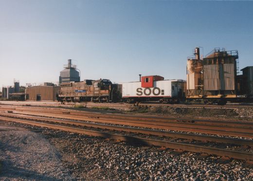 A SOO Line diesel locomotive and caboose are parked on parallel tracks in a sunny rail yard, with industrial buildings and a Mannheim station sign in the background.